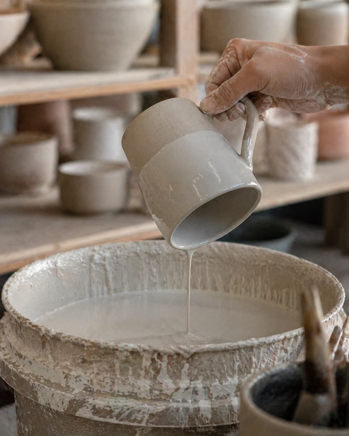 A clay-dusted hand tipping a stoneware mug to pour pale glaze in a thin stream into a tall studio bucket, drips and runs frozen mid-air, bisqued forms on shelves softly out of focus behind.