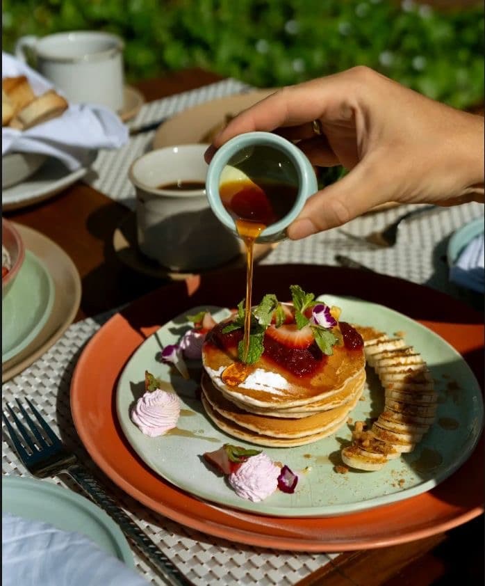 Syrup poured from a small ceramic pitcher onto a stack of pancakes, plated on hand-thrown stoneware in a sunlit garden setting.