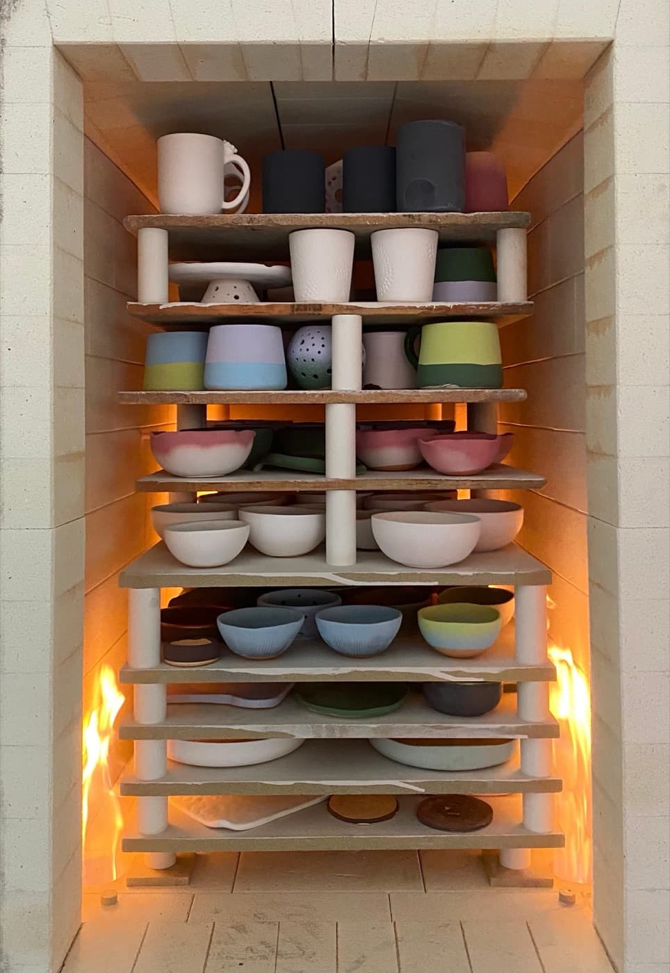 Gas kiln stacked floor-to-ceiling with bisqued and glazed pieces: mugs, cups, bowls, and platters arranged on kiln shelves with flame visible at the base of the chamber.