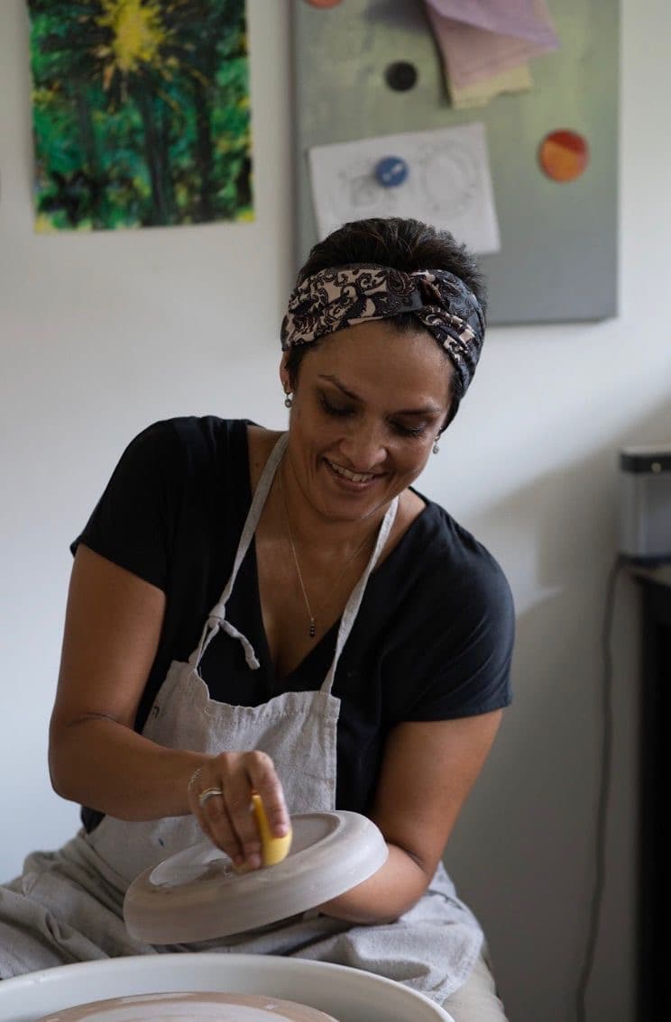 Mihiri de Silva at her studio wheel: paisley headscarf, white apron over a black tee, smiling as she sponges the rim of a freshly thrown plate.