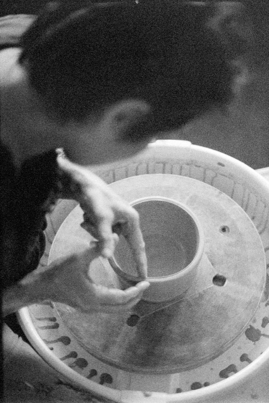 Black-and-white film photograph looking down at a potter's wheel: two hands shaping the wall of a vessel as the wheel head spins, the potter's silhouette dark above the rim.
