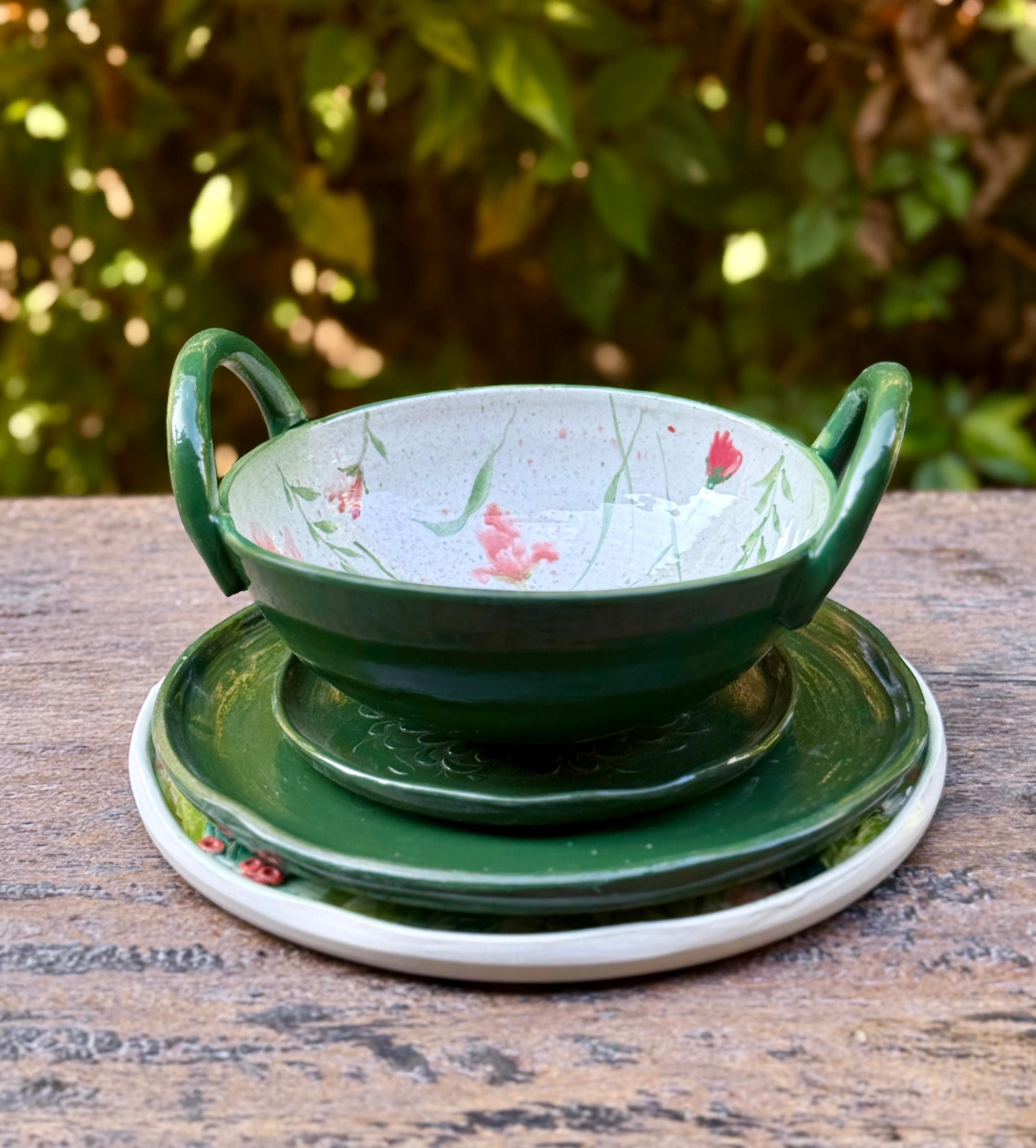 A green-glazed stoneware bowl with twin handles, hand-painted with red poppies inside, resting on two green plates atop weathered wood with garden foliage behind.