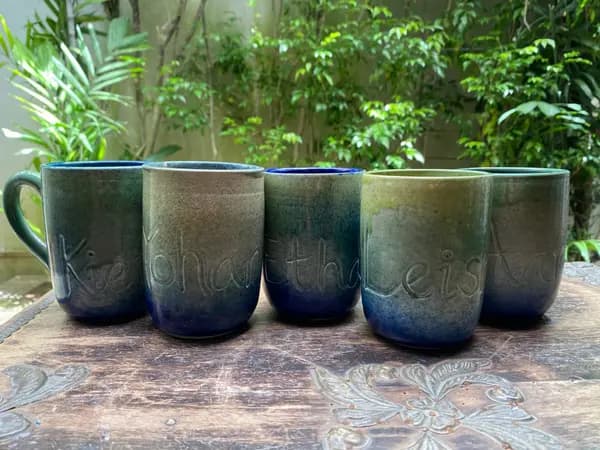 Five ceramic mugs with names engraved, featuring blue and green gradient glazes on a carved wooden table.