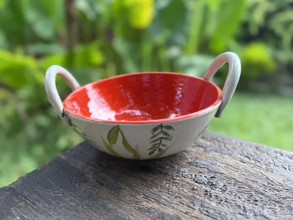 Ceramic bowl with vibrant orange interior, white exterior with green leaf motifs, and two handles on a wooden surface.