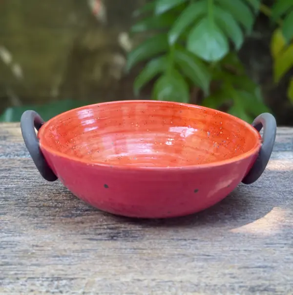 Vibrant pink and orange glazed ceramic thaachiya bowl with two black side handles on a wooden surface.