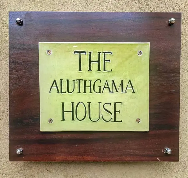 Square light green ceramic sign reading 'THE ALUTHGAMA HOUSE' mounted on dark wood with silver hardware.