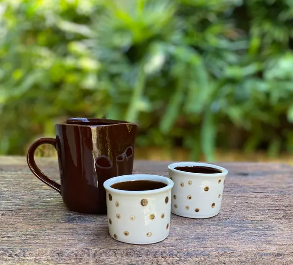 Dark brown ceramic jug and two white polka-dotted cups on a wooden table with a lush green background.