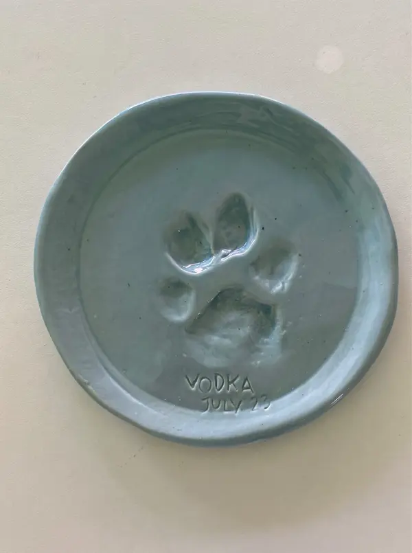 Light blue glazed ceramic dish with a paw print impression and the inscription 'VODKA JULY 23'.