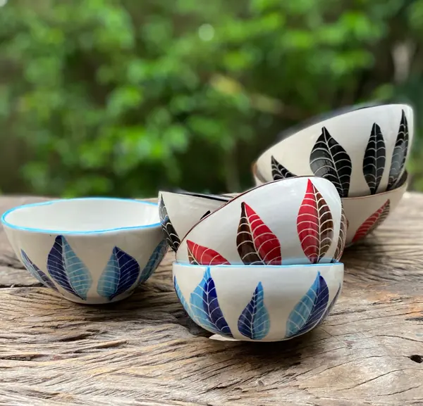 Hand-painted ceramic bowls with colorful leaf patterns in blue, red, and black on a rustic wooden table.