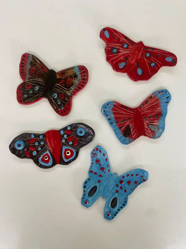 Five handmade ceramic butterflies in red, blue, and black glazes with intricate dotted patterns on a white surface.