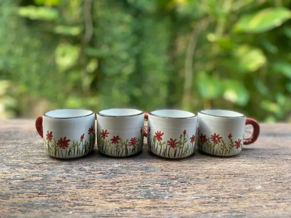 Four small white ceramic mugs with hand-painted red flowers and red handles on a wooden table.