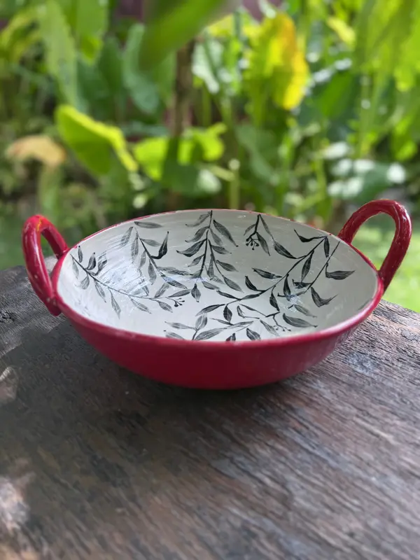 Red ceramic bowl with two handles and a white interior featuring a black leaf pattern on a wooden surface.