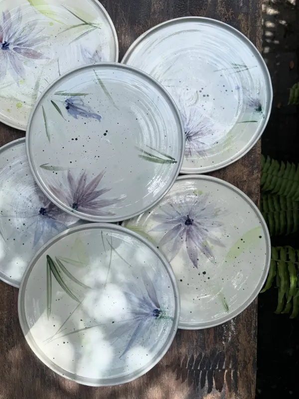 Five round ceramic plates featuring hand-painted purple flowers and green leaves on a speckled light gray glaze.