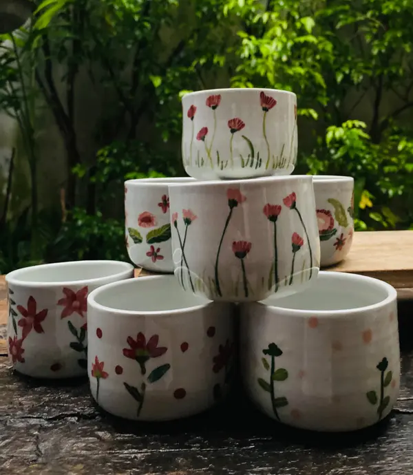 Stacked white ceramic cups with hand-painted red and pink flowers against a lush green garden backdrop.