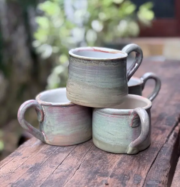 Four handmade ceramic mugs with muted sage, pink, and purple glazes stacked on a rustic wooden board.