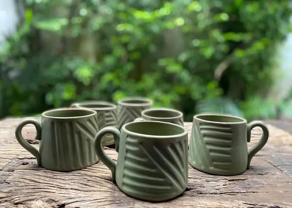 Six sage green matte mugs with carved geometric patterns on a weathered wooden surface with a lush green background.