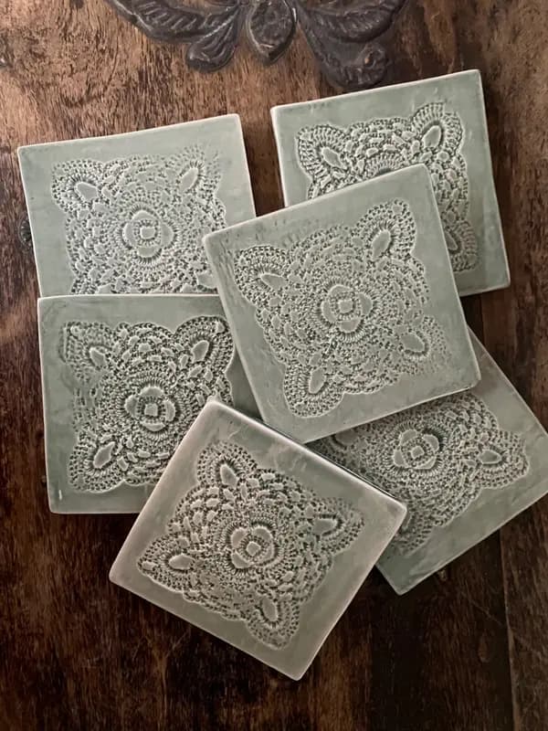 Square sage green ceramic coasters with embossed lace doily patterns, arranged on a dark wood surface.