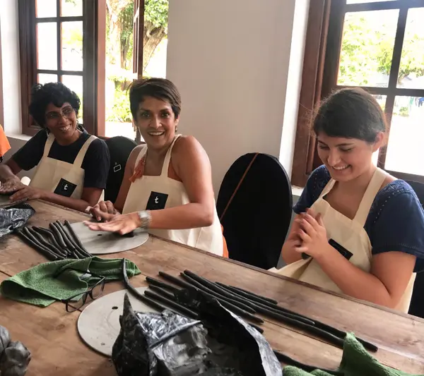Three women in white aprons smile while rolling dark clay coils on a wooden table during a pottery workshop.