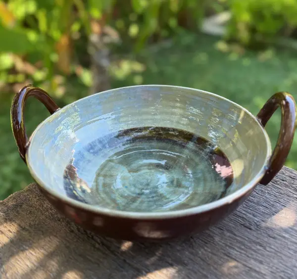 Ceramic thaachiya bowl with side handles, featuring an iridescent blue interior and a dark brown glazed exterior.