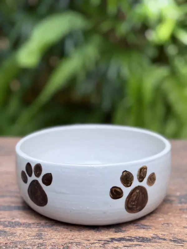 White ceramic pet bowl with hand-painted brown paw prints on a wooden surface with a garden background.