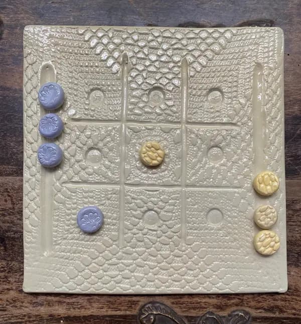 Beige textured ceramic tic-tac-toe board with small lilac and yellow game pieces on a dark wood surface.