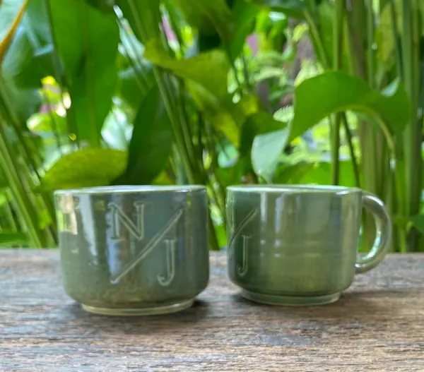 Two sage green ceramic mugs with engraved monograms on a wooden surface with a lush green garden background.