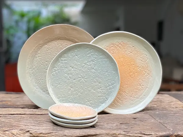 Three textured ceramic plates and a stack of small dishes with intricate embossed patterns in sage, beige, and coral.