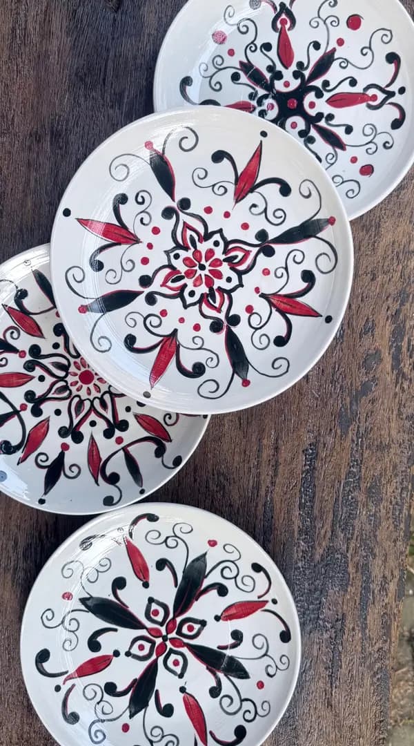 Four white ceramic plates with hand-painted red and black mandala patterns displayed on a dark wooden surface.