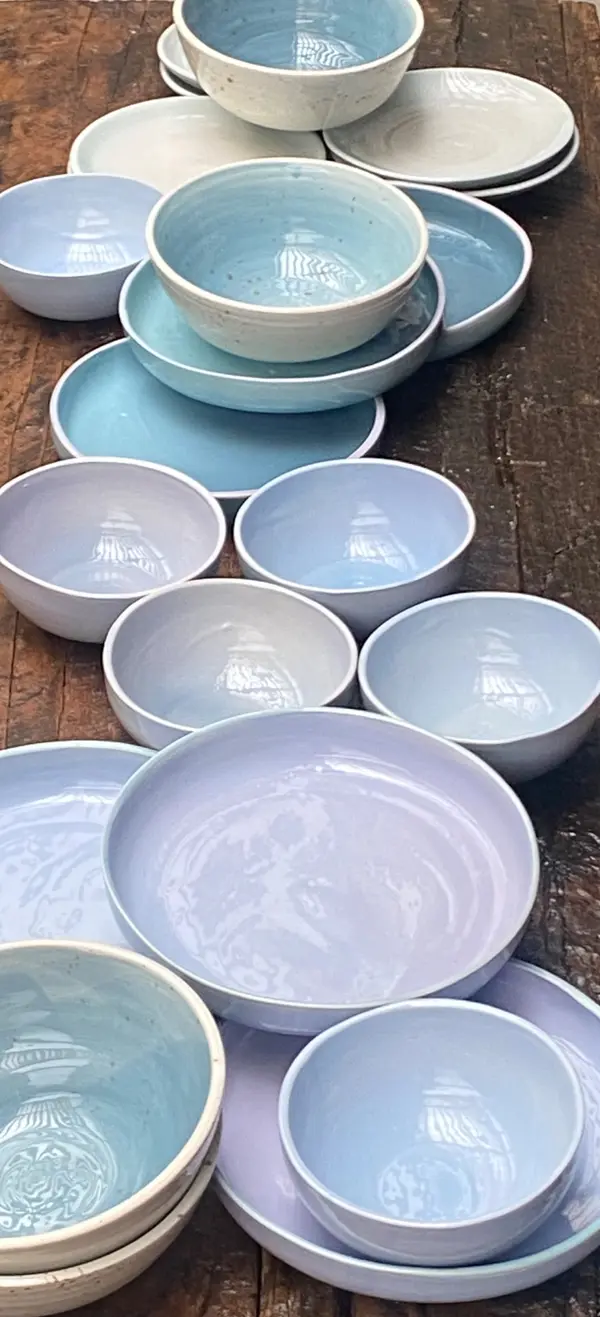 Handmade ceramic bowls and plates with light blue and lilac glazes arranged on a wooden surface.