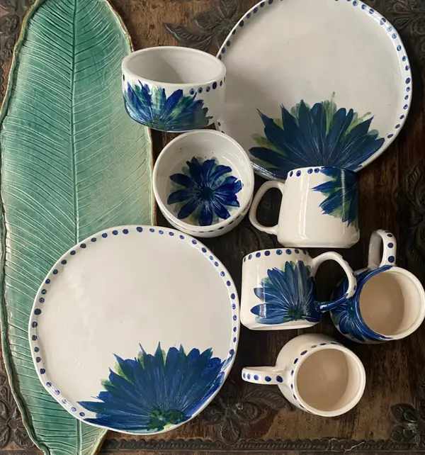 White ceramic mugs, bowls, and plates with blue floral art and a large green leaf platter on wood.