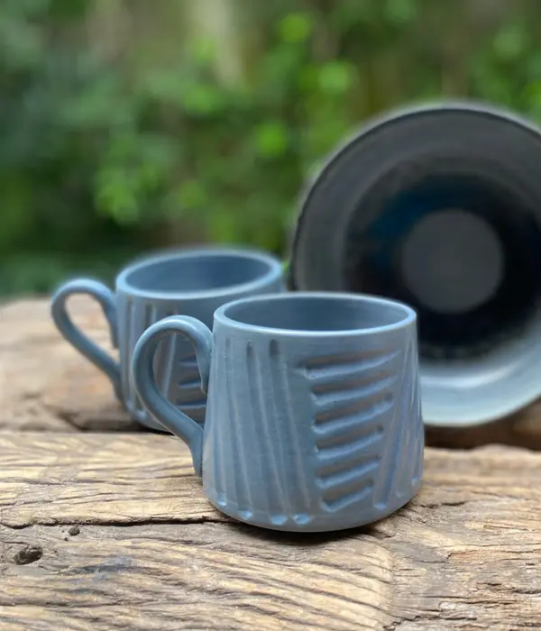 Two light blue-gray mugs with carved geometric patterns on a rustic wooden surface with a green background.
