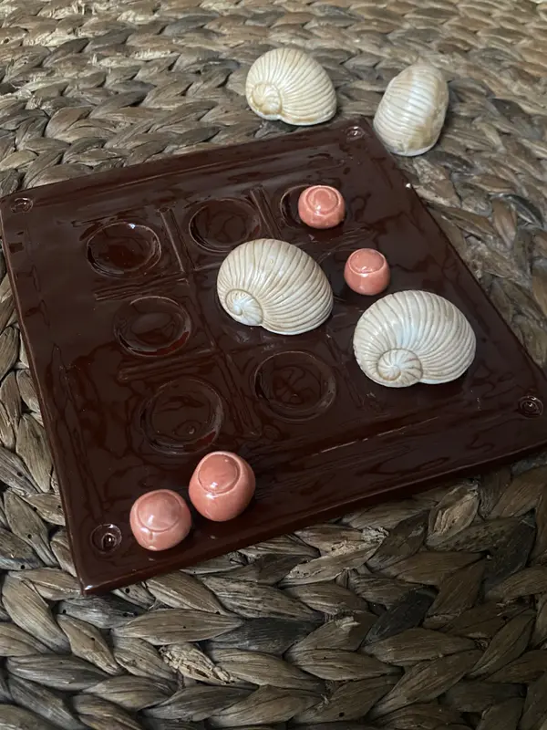 Ceramic tic-tac-toe board in glossy dark brown with snail shell and coral sphere game pieces on a woven mat.