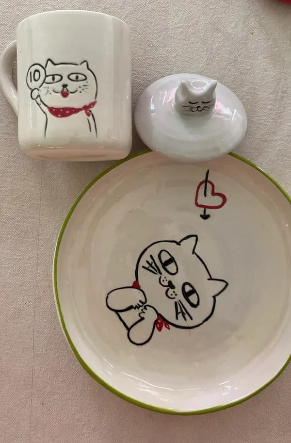White ceramic mug and plate set featuring hand-drawn black cat illustrations with red and green accents.