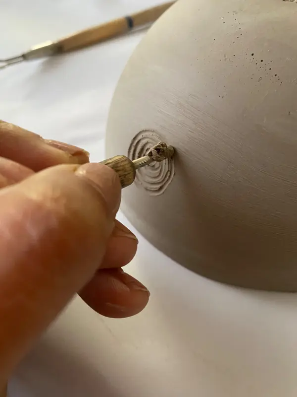 Close-up of a hand carving a spiral pattern into a raw beige clay bowl using a fine metal tool.