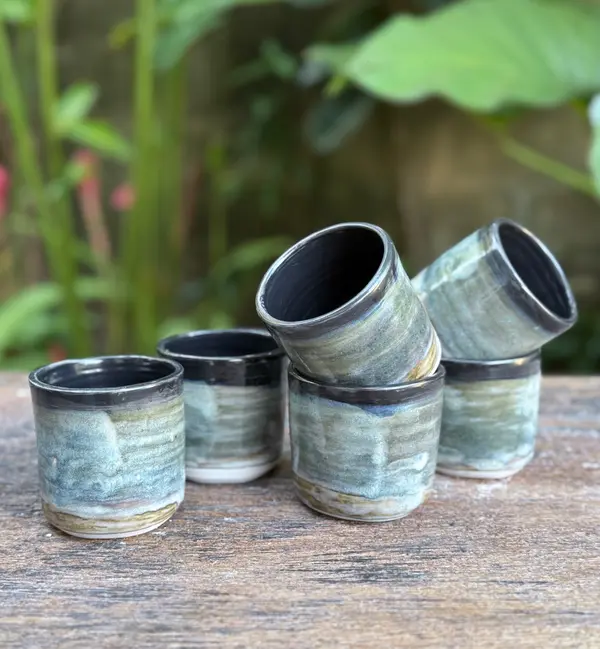 Five handleless ceramic cups with blue-green variegated glaze and dark rims stacked on a wooden surface.