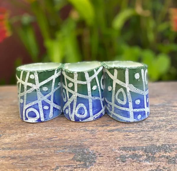Three small pottery cups with green and blue ombre glaze and white geometric patterns on a weathered wood table.