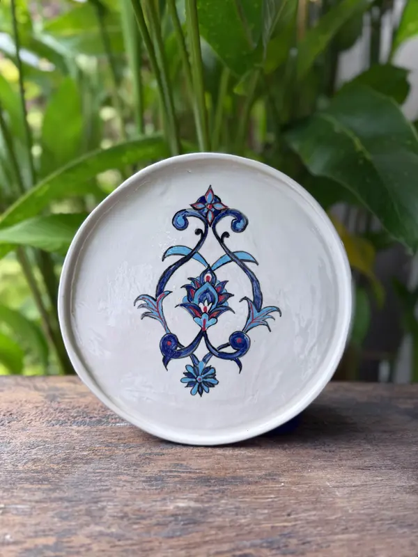 Circular white ceramic plate with a symmetrical hand-painted blue floral design on a wooden surface with plants.