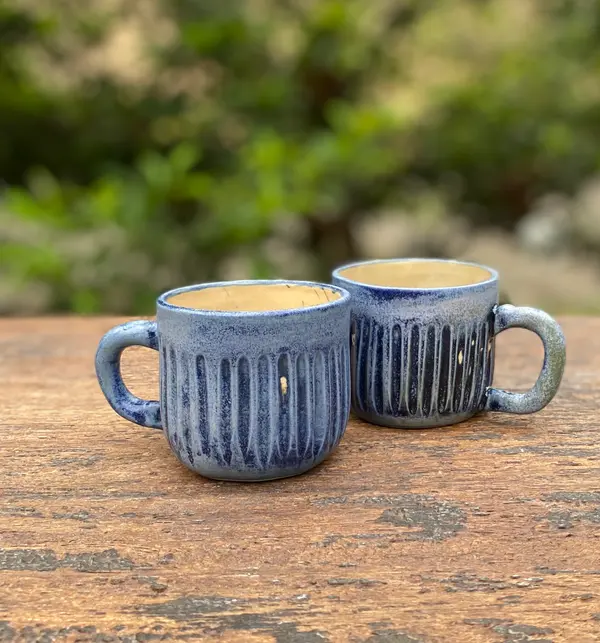Two light blue ceramic mugs with vertical fluted carvings on a weathered wooden table with a blurred green background.