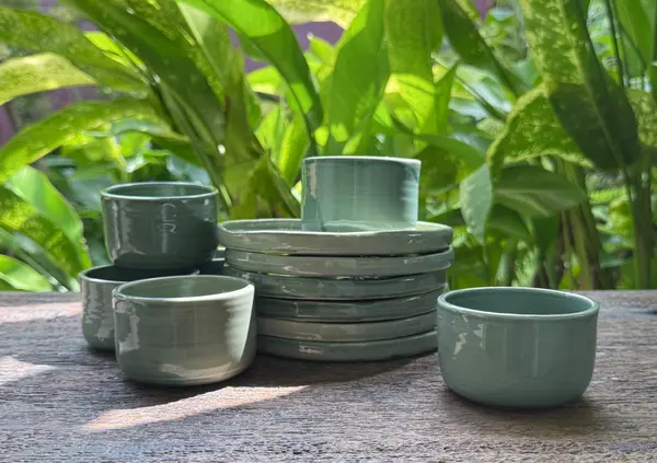 A set of glossy sage green ceramic bowls and a stack of matching small plates on a wooden table with green foliage.