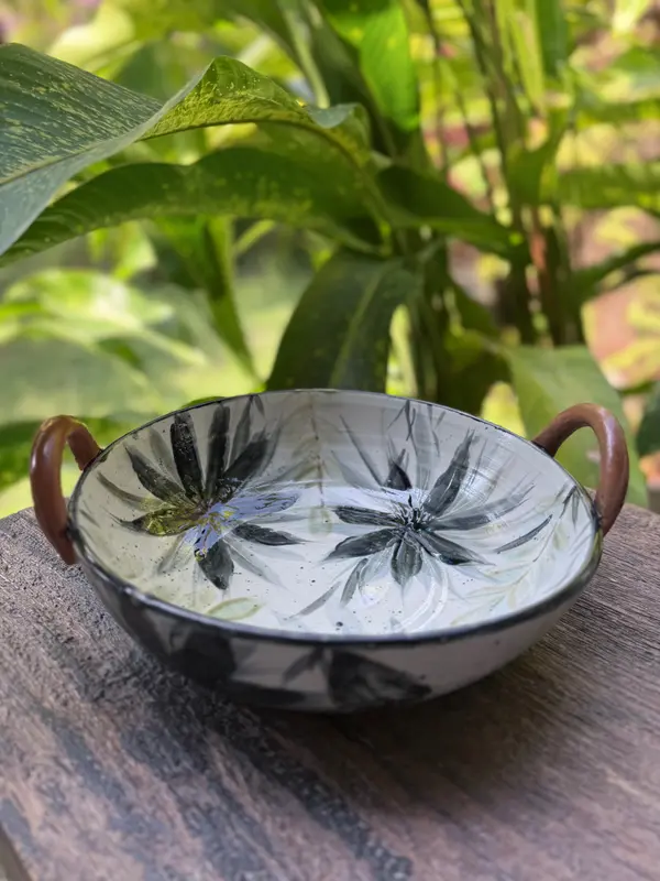 Hand-painted white ceramic bowl with black floral patterns and brown side handles on a rustic wooden surface.