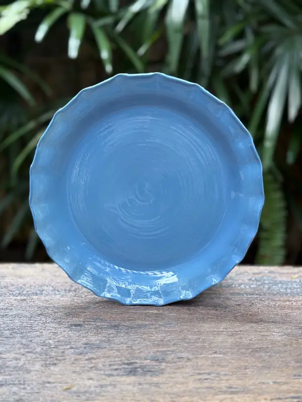 Glossy light blue ceramic plate with scalloped edges standing on a wooden surface against a leafy background.