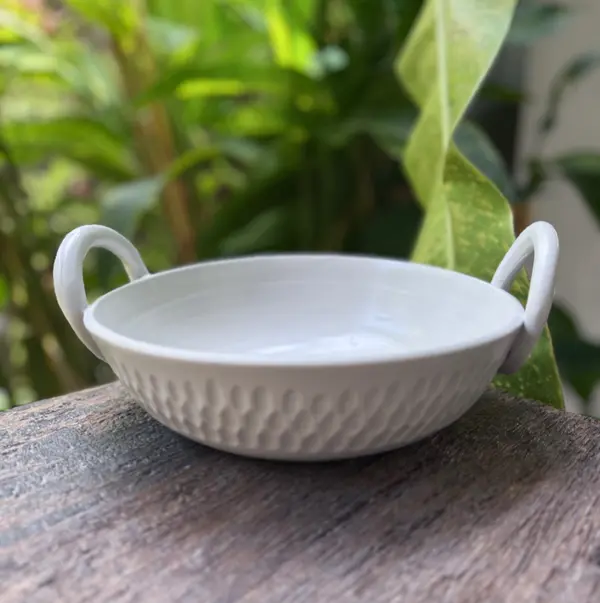 White ceramic thaachiya bowl with two handles and a dimpled textured exterior sitting on a wooden surface.