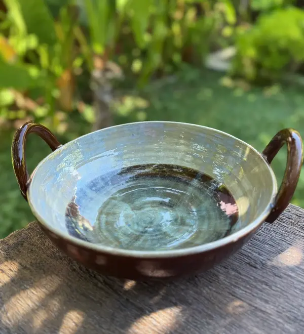 Handmade ceramic Thaachiya bowl with twin handles, light blue interior glaze, and dark brown exterior on wood.