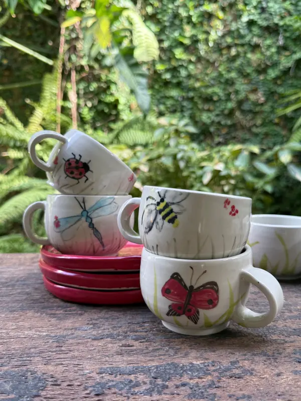Hand-painted white ceramic mugs with insect motifs and red saucers on a wooden table outdoors.