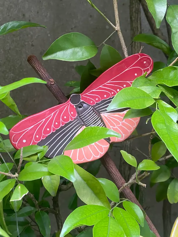 Ceramic cicada sculpture with red and black patterned wings, perched on a branch among green leaves.