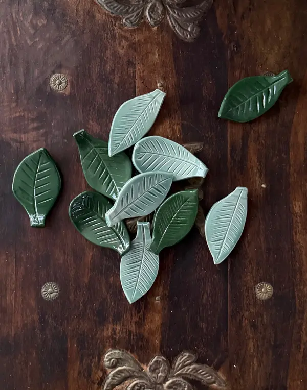Small glazed ceramic leaves in dark green and sage with carved veins, scattered on a dark wooden surface.