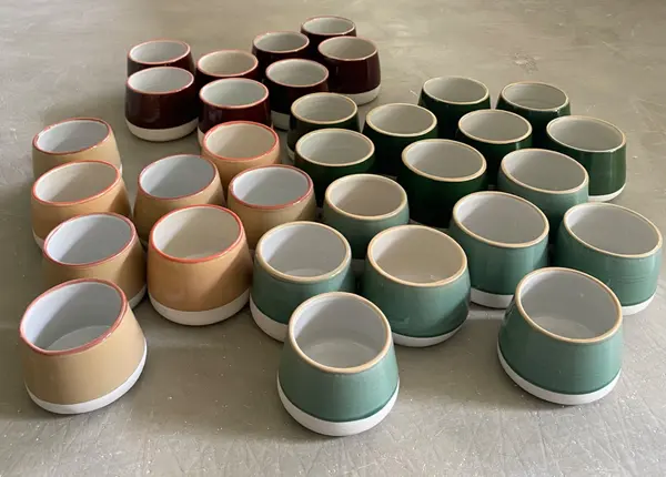 Array of ceramic handle-less cups in sage, dark green, and tan earth tones on a light gray surface.
