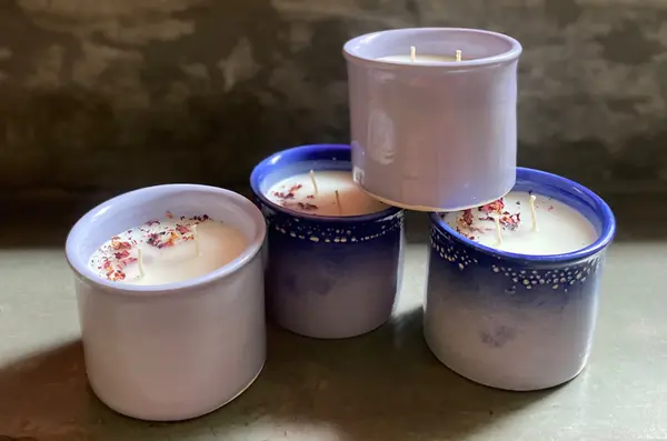 Four cylindrical ceramic candles in shades of lilac and cobalt blue with dried flower petals on top.