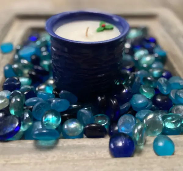 Cobalt blue textured ceramic candle holder filled with white wax and holly, surrounded by blue glass pebbles.