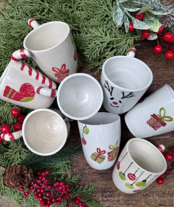 White ceramic holiday mugs with festive hand-painted motifs arranged on evergreen branches with red berries.